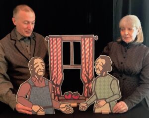 Two people, one man and one woman, both dressed in grey, each holding onto a 2-D wooden puppet of an older person who are standing in front of a set window, facing each other.